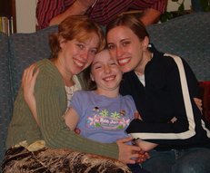 two big girls seated on the sofa, on either side of a small girl whom they are hugging
